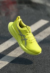 Baskets de course jaunes avec un dessus en maille léger, présentant des accents argentés et un design épuré, surélevées au-dessus d'une surface texturée.