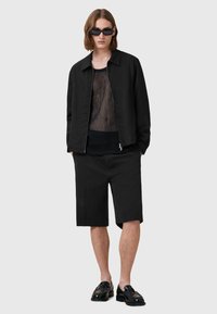 Schwarze Zip-Jacke über einem schwarzen Mesh-Top, kombiniert mit schwarzen Shorts bis zum Knie und glänzenden schwarzen Schuhen. Minimalistisches Design und Texturdetails.