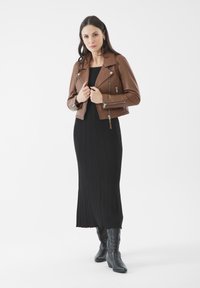 Giacca biker in pelle marrone con dettagli zip, indossata sopra un maxi dress nero a costine e stivali cowboy neri. Texture liscia, design corto.