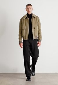 Olivgrüne Wollmischung-Jacke mit Kragen, zwei vorderen Taschen und Reißverschluss, kombiniert mit schwarzen Hosen und Schuhen, vor einem neutralen Hintergrund.