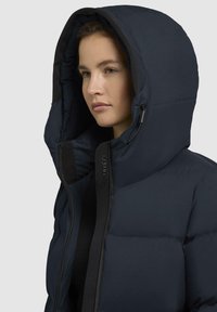 Giacca puffy blu navy con un grande cappuccio, texture liscia e chiusura con zip. Presenta un logo sulla parte frontale e cordini regolabili.