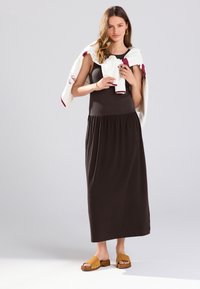 Longue robe brune avec un corsage ajusté et une jupe froncée, associée à un pull crème drapé sur les épaules et des sabots bruns.