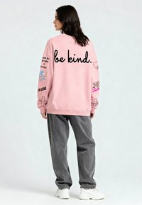 Rózsaszín pulóver, amelyen a "legyél kedves" felirat található, színes grafikákkal az ujjakon. Szürke, laza szárú nadrággal és fehér sportcipővel párosítva.