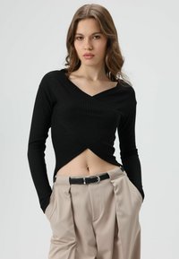 Zwart geribd topje met lange mouwen, een V-hals en een overslagontwerp, gecombineerd met beige hoge taillebroeken en een zwarte riem met zilverkleurige gespen.