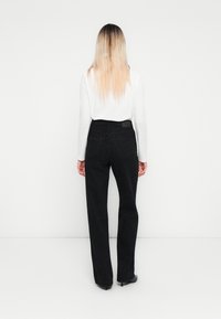 Person with long blonde hair wearing a white long-sleeve top and high-waisted black pants with a subtle leopard pattern, standing facing away.