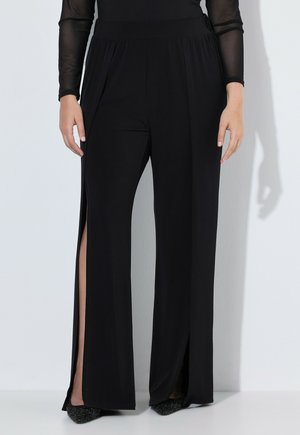 Pantalon noir à jambes larges avec taille haute et fentes latérales dévoilant les jambes, porté avec des chaussures noires et un top transparent à manches longues.