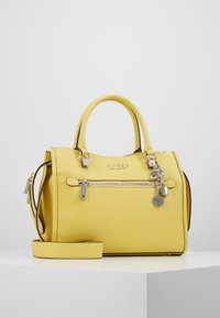 Keltainen kuvioitu nahkainen Guess-laukku, jossa on kaksinkertaiset kahvat, etupuolella vetoketjutasku, hopeinen logo ja avaimenperäkoristeet valkoisella esittelypinnalla.