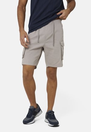 Mężczyzna w jasnoszarych szortach cargo ze ściągaczem w pasie oraz granatowymi sneakersami, lekko podnoszący granatową koszulkę.