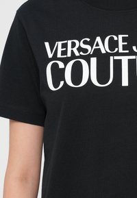 Juodas medvilnės marškinėlis su dideliu baltu tekstu, kuris ant priekinės dalies skaito "VERSACE JEAN COUTURE". Trumpomis rankovėmis ir apvaliu kaklu.