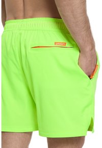 Neon žalios spalvos maudymosi šortai iš lengvos medžiagos, turintys elastingą juosmenį, šonines kišenes ir oranžinį užtrauktuko akcentą nugaros pusėje.
