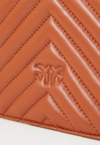 Browns ādas maku ar quiltētu zigzaga rakstu un iegrebtu logotipa detaļu. Iezīmēts ar teksturētu virsmu un kontrastējošām izšuvēm.