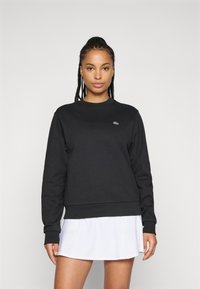 Svart hettegenser laget av bomullsblanding, med rund hals, lange ermer og en liten grønn Lacoste-logo på brystet.