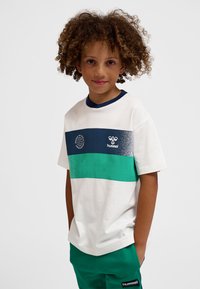 Göndör hajú gyerek fehér pólóban, kék és zöld csíkokkal, zöld nadrágban, zsebébe dugott kézzel áll egy egyszínű háttér előtt.