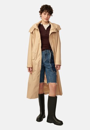 Un imperméable beige avec un col haut, une fermeture à boutons et des poches latérales, associé à un short en jean déchiré et des bottes en caoutchouc noires.