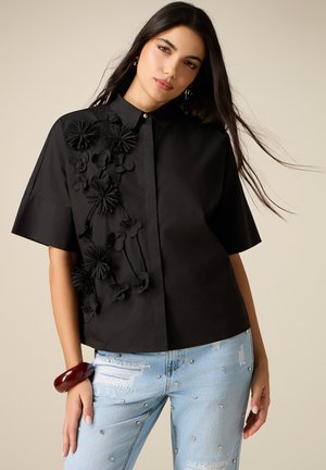 Femme aux longs cheveux foncés portant un t-shirt noir à manches courtes avec des applications florales et un jean bleu clair déchiré orné de strass.