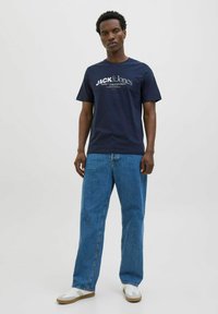 Marineblå bomulds T-shirt med hvid logo tekst, parret med løse, lyseblå denim jeans og hvide sneakers med beige accenter.