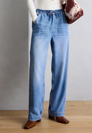 Personne portant un jean large bleu clair avec taille à cordon, un haut blanc à manches longues, des chaussures marron à bout carré, et tenant un sac marron et blanc.