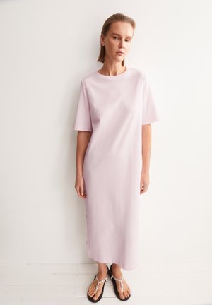 Vrouw staat tegen een witte muur en draagt een lichtroze midijurk met korte mouwen en zwarte sandalen met witte bandjes.