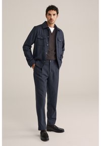 Bărbat îmbrăcat într-o jachetă și pantaloni bleumarin, cu un pulover cu guler în V de culoare maro închis și mocasini negri, stând în fața unui fundal bej.