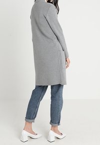 Grauer langer Cardigan mit geradem Design, seitlichen Taschen und weicher Textur, kombiniert mit hochgekrempelten Jeans und weißen Blockabsatzschuhen.