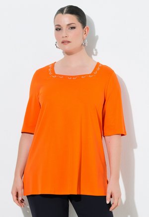 Donna con capelli scuri, indossa una maglietta arancione a maniche corte con scollo decorativo e orecchini a cerchio argentati, in piedi davanti a uno sfondo bianco.