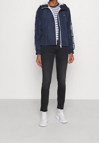 Person klädd i en marinblå jacka med huva och vit "Tommy Jeans" text på ärmarna, grå randig tröja, svarta jeans och vita sneakers på grått golv.