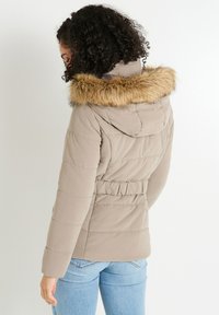Chaqueta de abrigo beige con capucha forrada de piel sintética, cinturilla elástica y patrón de acolchado, combinada con jeans de color azul claro y un fondo liso.