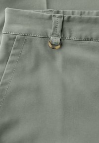 Gros plan sur un pantalon en tissu gris clair montrant la ceinture, un passants avec un détail d'anneau beige, et la couture de la poche latérale.