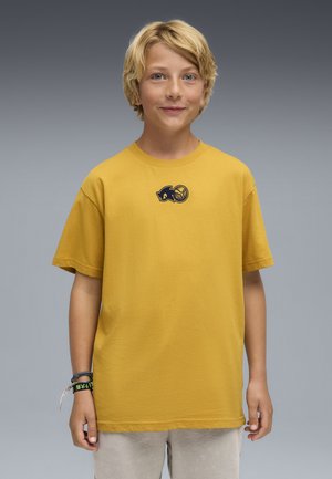 Garçon blond souriant portant un T-shirt jaune moutarde avec un petit logo noir et un short gris clair sur un fond gris neutre.