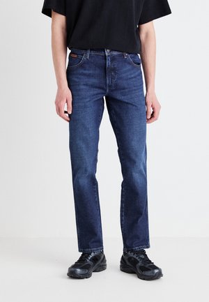 Dark blue denim jeans with a straight-leg cut, five-pocket design, and contrasting stitching, paired with black athletic shoes.
