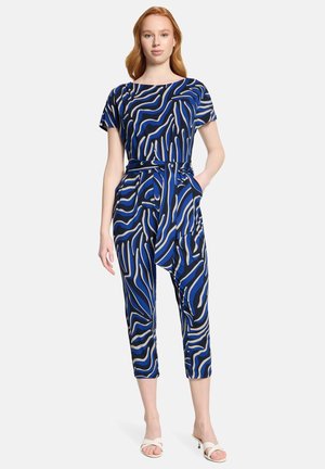 Kobieta ubrana w niebieski, czarny i beżowy kombinezon w abstrakcyjne paski, z krótkimi rękawami i paskiem przewiązanym w talii, w połączeniu z białymi sandałami na obcasie.