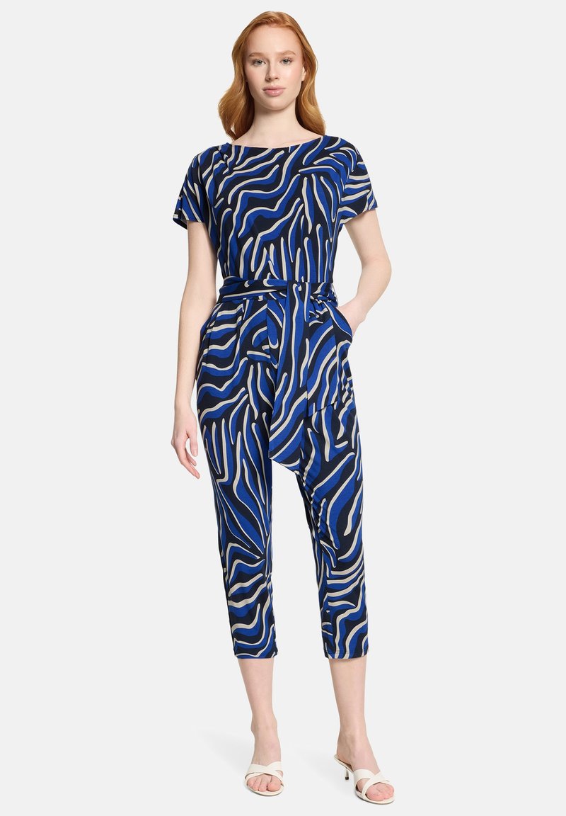 Femme portant une combinaison à manches courtes à rayures abstraites bleues, noires et beiges, avec une ceinture nouée à la taille, associée à des sandales blanches à talons.