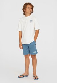 T-shirt blanche à manches courtes avec logo, shorts bleus et tongs bleues. Coupe décontractée avec un style relax. Tissu lisse avec des détails subtils.