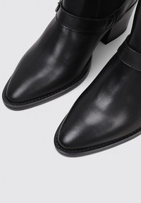 Bottes noires en cuir à talon aiguille et bout pointu. Elles présentent une sangle décorative autour de la cheville et une finition lisse et brillante.