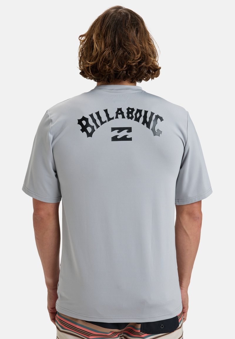 Camisa de manga corta gris claro, con un gran logo negro de "Billabong" acompañado de un gráfico de ola. Textura suave y corte relajado.