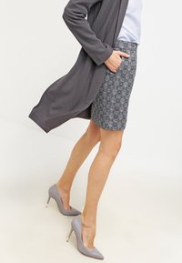 Cardigan grigio sopra una gonna aderente con un motivo in bianco e nero. Tacchi alti grigi completano l'abbigliamento. Tessuti lisci e design sartoriale.
