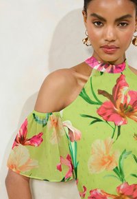 Blusa floreale verde con maniche senza spalline, caratterizzata da motivi floreali rosa e arancioni, tessuto strutturato e un collo alto con bordatura rosa.