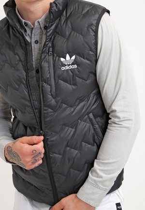 Zwarte geweven puffer vest met een ritssluiting aan de voorkant, hoge kraag en Adidas-logo. Draagt over een lichtgrijze lange mouw shirt.