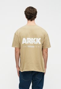 Muž v béžovém oversized tričku s bílým textem "ARKK" na zadní straně, v kombinaci s tmavě modrými džíny, stojící na čistém bílém pozadí.