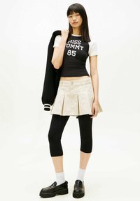 Zwart-wit cropped top met de tekst "MISS TOMMY 85", beige geplooide rok, zwarte legging, witte sokken en zwarte plateauzolen.
