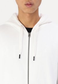 Witte rits hoodie met een trekkoordcapuchon, met een soepele textuur, en een klein logo op de borst. Ritssluiting heeft een zilveren afwerking.