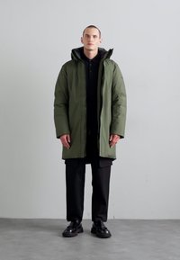 Long green puffer coat with a high collar, full zip, and hood, worn over a black polo shirt and black pants, paired with glossy shoes.