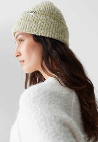 Zielony, textured beanie o splocie, z zagiętym brzegiem i czarną metką z logo. Noszony z białym, puszystym swetrem, co podkreśla miękki, ciepły wygląd.