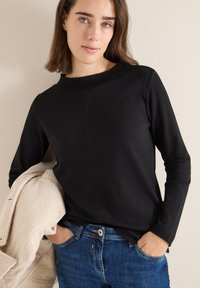 Black long-sleeve top with a crew neckline, featuring a smooth texture and seam detailing, paired with blue denim jeans and a beige outer layer.