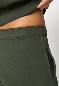 Pantaloni joggeri din bumbac verde închis, cu talie elastică, având buzunare laterale și o textură netedă, parțial acoperiți de un top asortat.