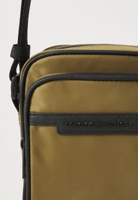 Borsa a tracolla in nylon marrone con finiture nere, dotata di una tasca frontale con zip e di un'etichetta nera marchiata con "TOMMY HILFIGER".