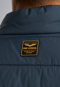 Marinblå jacka med en quiltad textur, med ett rektangulärt gult och svart märke, inskrivet "PME LEGEND" och "TESTED AT HIGH ALTITUDE."