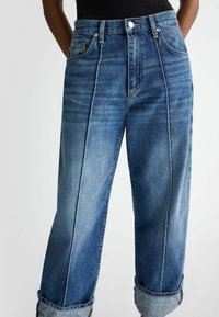 Jeans de denim de peso pesado en un lavado azul medio, con un diseño de pierna recta, bolsillos delanteros y dobladillos rematados. Costuras en contraste visibles.