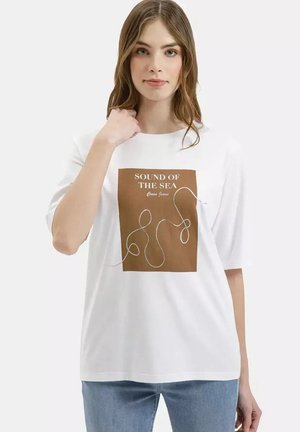 Młoda kobieta w białej koszulce z brązowym kwadratowym nadrukiem i napisem "SOUND OF THE SEA" oraz jasnoniebieskich dżinsach, stojąca na białym tle.