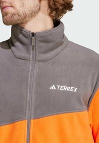 Fleecejakke i grå med et orange nederste panel. Har høj krave og en frontlynlås. Vist logo: "TERREX" i hvid.